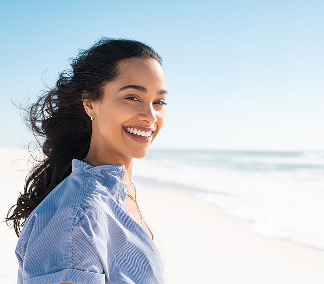 Smiling Latina woman standing on the beach with the wind in her hair, representing empowerment, healing, and culturally responsive counseling.