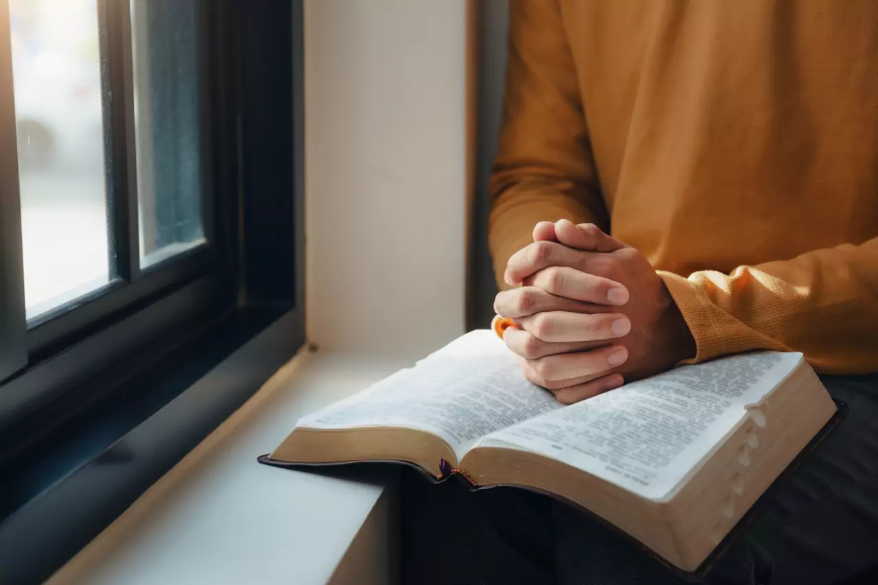 Close-up of hands gently folded over a Bible near a window.